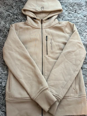Beige hoodie från Sail Racing - Snygg beige hoodie från Sail Racing med dragkedja framtill, huva och en bröstficka med dragkedja. Tillverkad i mjukt material och har ribbade muddar vid ärmslut och nederkant. Perfekt för en avslappnad och stilren look. Vid luvan har den fått två fläckar men det går bort av att tvätta 