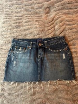 Blå jeanskjol med fransig kant - Snygg blå jeanskjol i kort modell med fransig, rå nederkant och slitna detaljer framtill. Klassisk femficksdesign och dekorativa bakfickor med knappar.osäker på storlek men skulle gissa på S skrev för mer mått annars!
