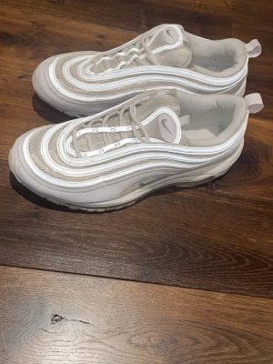 Nike Air Max 97 vita sneakers - Nike Air Max 97 sneakers i vitt och ljusgrått med ikoniska vågformade linjer och synlig Air-sula. Ovandelen är i mesh och syntetmaterial, med reflekterande detaljer och klassisk Nike Swoosh på sidan och plösen. Skorna har snörning och dragflik bak.