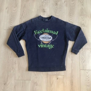 Collagetröja vintage stil - Snygg sweatshirt med vintage inspirerad design. Framsidan täcks av ett tryck i sportig stil och med en sliten finish. Texten i maskinbroderi säger Reclaimed vintage och är även märket på plagget. Tröjan är mörkblå med en flammig urtvättad look. 100% bomull. Passar storlek xs för en avslappnad passform. 