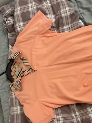 Persikofärgad Burberry skjorta M - Snygg persikofärgad skjorta från Burberry med klassisk rutig krage och knappslå. Skjortan har korta ärmar och är tillverkad i bomull, perfekt för en trendig och avslappnad look.