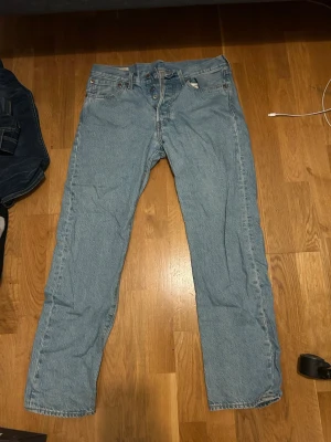 Levi's 501 ljusblå raka jeans - Klassiska Levi's 501 jeans i ljusblå denim med rak passform och knappgylf. Jeansen har fem fickor, tydlig Levi's-lapp bak i midjan och röd etikett på bakfickan. Perfekta för en avslappnad och tidlös stil.