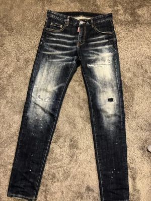 Mörkblå slitna jeans från Dsquared2 - Säljer ett par mörkblå jeans från Dsquared2 med skinny fit och slitningar på knäna samt distinkta tvättade detaljer. Jeansen har klassisk femficksmodell, vita färgstänk och patch bak i läderimitation. Perfekta för en avslappnad streetstil.