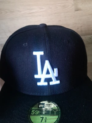 Svart LA-keps från New Era - Säljer en svart fitted keps från New Era med klassisk LA-logga broderad i vitt framtill. Kepsen är modellen 59FIFTY och har en böjd skärm samt MLB-logga baktill. Perfekt för dig som gillar streetwear och sportig stil.