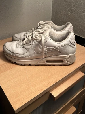 Nike Air Max 90 vita sneakers - Klassiska Nike Air Max 90 sneakers i helvitt läder med mesh-detaljer. Skorna har den ikoniska synliga Air Max-sulan och en chunky siluett. Perfekta för dig som gillar streetwear och sportig stil.