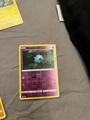 Pokémonkort Woobat holografisk - Säljer ett Pokémonkort med Woobat-motiv. Kortet har en holografisk lila bakgrund och gul ram. Text och symboler är tryckta i svart och vitt. Perfekt för samlare eller Pokémonfans som vill utöka sin kortsamling.