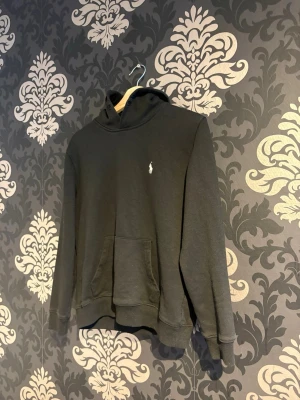 Svart hoodie från Polo Ralph Lauren - Svart hoodie från Polo Ralph Lauren med klassisk huva och känguruficka framtill. Liten vit broderad logga på bröstet. Tillverkad i mjukt bomullsmaterial och har ribbade muddar vid ärmslut och nederkant. Perfekt för en avslappnad och stilren look.