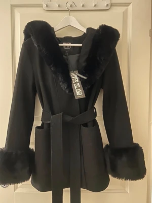 Svart kappa med fuskpäls från River Island - Stilren svart kappa från River Island med stora fickor, knytskärp i midjan och huva. Kappan har lyxiga detaljer i svart fuskpäls runt huva och ärmslut. Storlek 34. Nypris 899kr. 