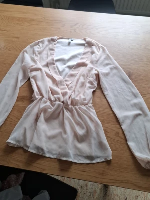 Ljusrosa omlottblus med v-ringning - Snygg ljusrosa omlottblus med djup v-ringning och resår i midjan som ger en fin siluett. Blusen har långa ärmar och är tillverkad i ett tunt, flowy material som känns luftigt och lätt.