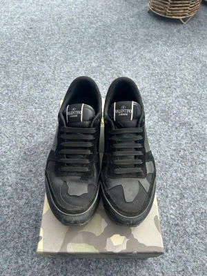 Svarta Valentino Garavani sneakers - Snygga svarta sneakers från Valentino Garavani med coola camo-detaljer och mix av mocka och läder. Skorna har lågt skaft, svarta snören och en robust sula med nitar på hälen. Perfekta för dig som gillar exklusiva och trendiga sneakers.