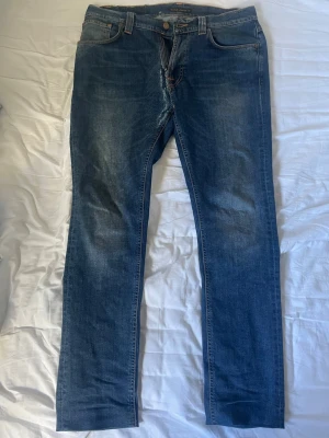 Blå Nudie Jeans - Snygga blå slim fit jeans från Nudie med klassisk femficksdesign och orange sömmar på bakfickan. Jeansen har en lätt tvättad look och är tillverkade i mjuk denim med stretch för extra komfort.