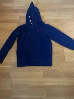 Marinblå hoodie från Polo Ralph Lauren - Snygg marinblå hoodie från Polo Ralph Lauren med klassisk huva och vit dragsko. Ikoniska röda loggan broderad på bröstet. Hoodien har känguruficka framtill och är gjord i mjukt bomullsmaterial. Nypris 2400. 