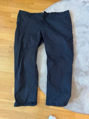Svarta chinos med rak passform - Säljer ett par svarta chinos med rak passform och klassisk design. Byxorna har bälteshällor, knapp och dragkedja fram samt fickor både fram och bak. Perfekta för en clean och stilren look. Materialet känns mjukt och bekvämt, troligen bomull eller bomullsblandning.