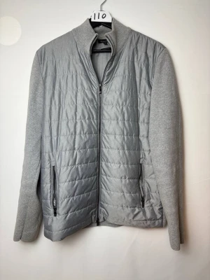 Massimo Dutti cardigan - Stilren ljusgrå cardiganjacka med quiltad framsida och släta stickade ärmar. Jackan har dragkedja framtill och två praktiska fickor med dragkedja. Perfekt för lager-på-lager och enkel att matcha med olika outfits. Plagg nmr 110