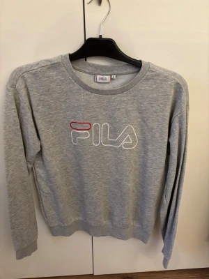 Grå sweatshirt från FILA - Snygg grå sweatshirt från FILA med klassisk logga i vitt och rött framtill. Tröjan har rund halsringning, långa ärmar och ribbade muddar vid ärmslut och nederkant. Perfekt för en avslappnad och sportig stil.