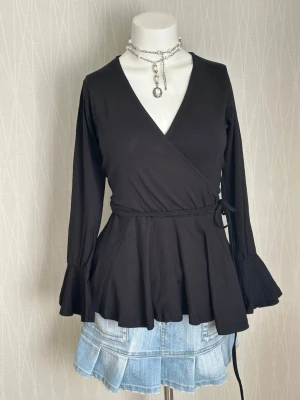 Svart omlottopp med volangärm - Svart omlottopp med långa ärmar och volang vid ärmslut. Toppen har knyt i midjan och v-ringad hals. Tillverkad i mjuk bomull, perfekt för en trendig och bekväm look.