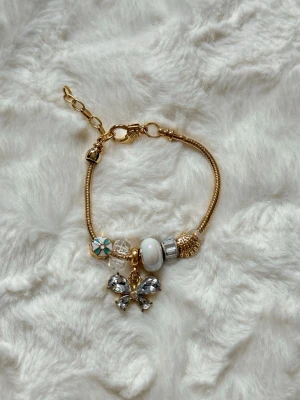 Guldfärgat armband med berlocker - Snyggt guldfärgat armband med flera berlocker, bland annat en fjäril med glittrande stenar, pärlliknande detalj, blommönster och dekorativa pärlor. Justerbar kedja och mix av guld, vitt och silverfärgade inslag. Perfekt accessoar för att lyfta din stil.