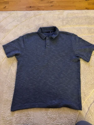 Blå pikétröja med krage - Snygg blå pikétröja med klassisk krage och två knappar framtill. Tröjan har korta ärmar och en lätt melerad look. Perfekt för en avslappnad men ändå stilren outfit. Från lager 157