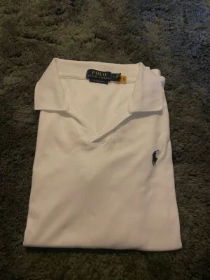 Vit pikétröja från Polo Ralph Lauren - Klassisk vit pikétröja från Polo Ralph Lauren med korta ärmar och den ikoniska broderade loggan på bröstet. Tröjan har en mjuk krage och är tillverkad i bomull för en bekväm känsla. Perfekt till jeans eller shorts för en clean look.