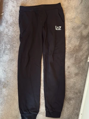 Svarta mjukisbyxor från EA7 160 Armani  - Svarta mjukisbyxor från EA7 Emporio Armani med smal passform och resår i midjan. Byxorna har fickor på sidorna och en vit EA7-logga tryckt på vänster lår. Perfekta för en avslappnad och sportig stil.