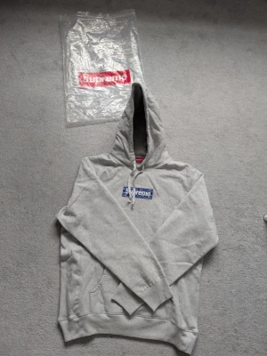 Grå Supreme hoodie med blå box logo - Supreme hoodie i ljusgrått med klassisk blå box logo på bröstet. Storlek L/XL. Hoodien har huva med dragsko, känguruficka och ribbade muddar. Perfekt för dig som gillar streetwear och vill sticka ut med en ikonisk look. 