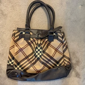 Rutig handväska med bruna detaljer - Snygg handväska från liknande burberry med klassiskt rutigt mönster i beige, brunt och svart. Väskan har dubbla handtag och detaljer i mörkbrunt läder samt dekorativt bälte upptill. Perfekt för dig som gillar tidlös och trendig design.
