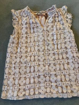 Vit spetsblus med volangdetaljer - Supersöt vit blus med broderad spets och volangdetaljer vid axlarna. Blusen är ärmlös och har v-ringning med liten knäppning i nacken. Perfekt för dig som gillar romantisk och feminin stil.