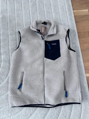 Beige fleeceväst från Patagonia - Mysig beige fleeceväst från Patagonia med marinblå detaljer. Västen har hög krage, två sidofickor med dragkedja och en bröstficka i marinblått tyg. Perfekt för lager-på-lager och har en sportig look med blå zippar.