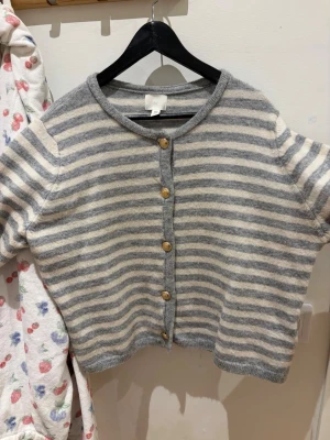 Randig gråvit kofta med träknappar - Mysig kofta med grå och vita horisontella ränder, rund halsringning och träfärgade knappar framtill. Koftan har en avslappnad passform och är stickad i ett mjukt material. Perfekt för lager på lager och enkel att styla till olika outfits.
