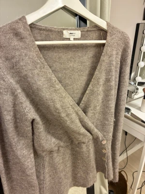 Beige omlottkofta från Selected Femme - Mysig beige omlottkofta från Selected Femme med v-ringning och knappar på sidan. Koftan har lång ärm och är stickad i ett mjukt material som känns skönt mot huden. Perfekt för lager på lager och enkel att styla till olika outfits.