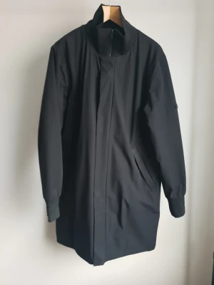 Svart minimalistisk vinterjacka med hög krage - Säljer en svart vinterjacka med minimalistisk design och hög krage. Jackan har dolda fickor framtill, dragkedja och knappslå. Materialet är vattenavvisande och har en slät finish. Perfekt för dig som gillar stilrena och funktionella plagg.