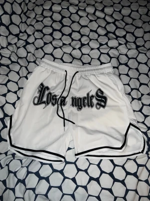 Vita Los Angeles shorts med snörning - Snygga vita shorts med svart text 'Los Angeles' framtill i gotisk stil. Shortsen har svart snörning i midjan och svarta detaljer längs kanterna. Perfekta för sommaren och har en sportig vibe.