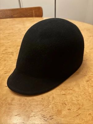 Svart filthatt från H&M - Säljer en svart filthatt från H&M i storlek M/56. Hatten är tillverkad i 100% ull och har en klassisk, rundad form med kort skärm. Insidan är dekorerad med ett svartvitt prickigt band för extra stilpoäng. En toppensnygg hatt!! Köpte och var svintaggad men den va för liten för mig :/ 100% ull