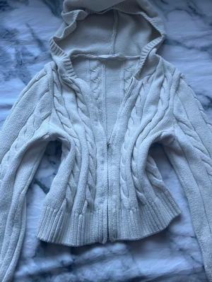 Vit kabelstickad hoodie med dragkedja - Säljer en vit kabelstickad kofta  med dragkedja. Perfekt för dig som gillar en mysig och stilren look. Materialet känns mjukt och stretchigt. Är i storlek 12y men är syd längre eftersom jag i storlek xs har haft den på väldigt stretchig och fin och passar bra med många outfits och använd bara några gånger men tyvärr inte passar min nya stil 