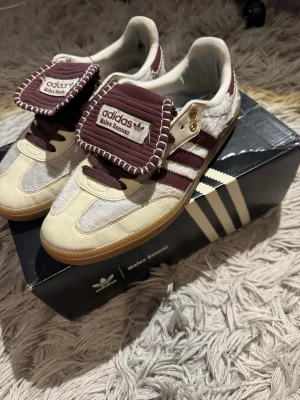 Adidas Wales Bonner skor - Unika Adidas Wales Bonner sneakers i beige och vinrött med klassiska tre ränder på sidan och grov gummisula. Skorna har en stor tunga med Wales Bonner-logga och detaljer i mocka och textil. Perfekta för dig som gillar retrostil och coola samarbeten. De säljs för ett sjukt bra pris pågrund av att de ör lite slitna som man ser på de sista bilderna. Som man ser på näst sista bilden så har aulan släppt lite men jag har försökt laga det genom att limma de. 