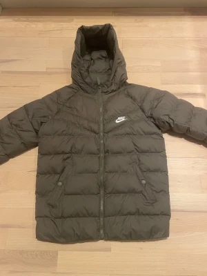 Olivgrön dunjacka från Nike - Snygg olivgrön dunjacka från Nike med huva och dragkedja framtill. Jackan har två snedställda fickor med knappar och ett vitt Nike-logotyptryck på bröstet. Perfekt för kalla vinterdagar och riktigt skön att bära. Kolla storleken på bilden, gissar att det motsvarar XS.