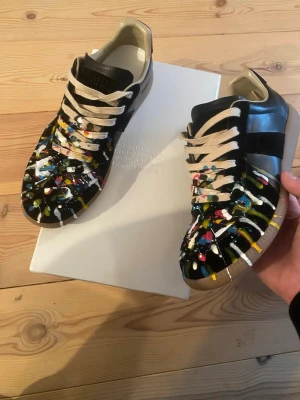 Maison Margiela Replica sneakers - Unika Maison Margiela Replica sneakers i svart läder med färgstänk i vitt, gult, blått, rött och grönt. Beigea skosnören och beige insida. Hör av dig vid fråger eller flera bilder tveka inte!