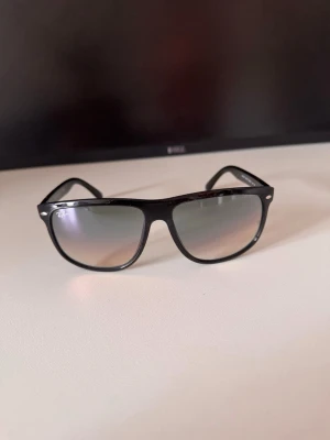 Svarta Ray-Ban solglasögon - Snygga svarta solglasögon från Ray-Ban med klassisk wayfarer-form. Bågarna är blanka och har Ray-Ban-loggan på sidan. Glasen är mörka och ger en cool look till alla outfits.