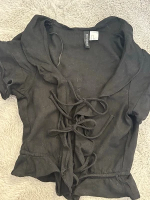 Svart snörad volang - Säljer en svart snörad volang topp från divided H&M i storlek XS. Toppen har en kort, öppen look men 3 knyten och volanger.  Den är använd två gånger!! Skriv till mig vid frågor eller intresse