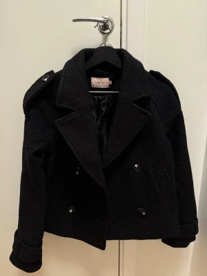Ny kort jacka  - Snygg kort svart ullrock från Nelly med dubbelknäppning, bred krage och axelklaffar. Rocken har två sidofickor och är fodrad för extra komfort. Perfekt för dig som vill ha en stilren och trendig look i höst och vinter.