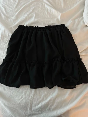 Svart volangkjol med resårmidja - Supersöt svart kortkjol med volangdetaljer och resår i midjan. Kjolen har en luftig och flowy känsla, perfekt för att styla med en crop top eller stickad tröja. Materialet känns lätt och ger kjolen ett snyggt fall.