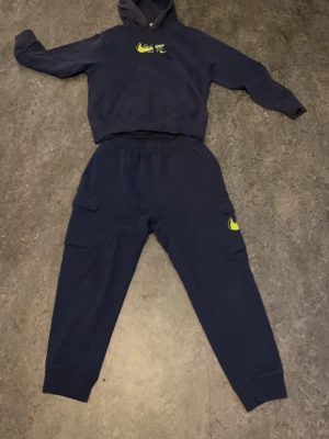 Mörkblå Nike hoodie med limegrön logga - Snygg mörkblå hoodie från Nike med limegrön logga på bröstet. Hoodien har en klassisk känguruficka framtill och justerbar huva. Tillverkad i mjukt material som känns skönt mot huden. Perfekt för dig som gillar streetwear och sportig stil.