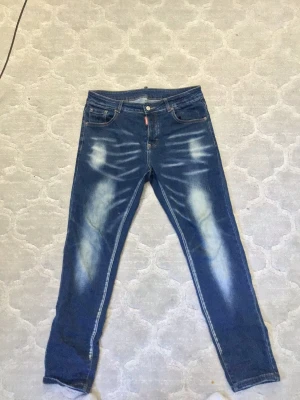 Blå jeans från Dsquared2 - Säljer ett par blå  jeans från Dsquared2 med slitna detaljer och ljusa tvättade partier framtill och baktill. Klassisk femficksmodell med knappgylf och Dsquared2-märke bak i linningen. Perfekta för en trendig och avslappnad look.