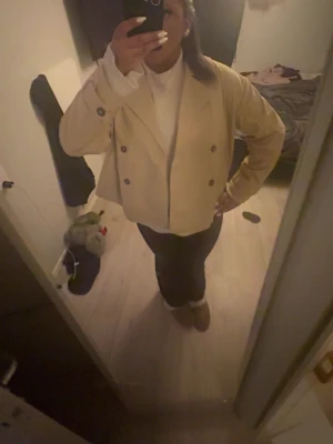 Beige dubbelknäppt kort rock - Stilren beige rock med dubbelknäppning och bred krage. Kortare modell som ger en modern look. Passar perfekt till både jeans och finare byxor. Inga synliga loggor eller märken. Troligtvis i ett mjukt tyg, eventuellt ullblandning.