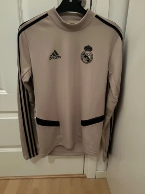 Beige långärmad Real Madrid tröja - Snygg beige långärmad träningströja från Adidas med Real Madrid-logga på bröstet. Tröjan har svarta detaljer, inklusive Adidas-logga, tre ränder längs ärmarna och svarta paneler vid fickorna. Perfekt för fotbollsträning eller chill.