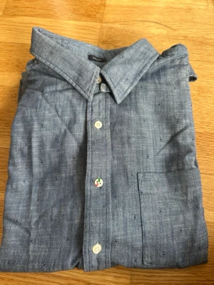 Blå skjorta från Boomerang, storlek L - Snygg blå skjorta från Boomerang i storlek L. Skjortan har klassisk krage, bröstficka och vita knappar framtill. Tillverkad i ett mjukt och svalt material som passar perfekt till många olika stilar.