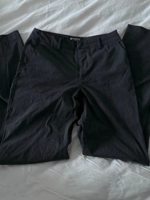 Randiga kostymbyxor från Madlady - Snygga svarta kostymbyxor från Madlady med diskreta vita kritstrecksränder. Byxorna har hög midja, hällor för skärp och raka ben. Perfekta för en stilren och trendig look. Materialet känns mjukt och följsamt.