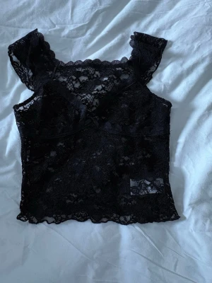 Svart spets topp - Snygg svart spets top  med breda axelband och vacker blommig spetsdesign. Transparent material och kortare modell som ger en trendig look. Perfekt att styla under en kavaj eller till jeans för en cool outfit.