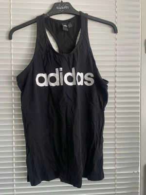 Svart linnetopp från Adidas - Säljer en svart linnetopp från Adidas med stort vitt logotryck framtill. Toppen har en sportig racerback-design och är tillverkad i mjuk bomull, perfekt för träning eller chill dagar. Enkel och clean look som funkar till det mesta.