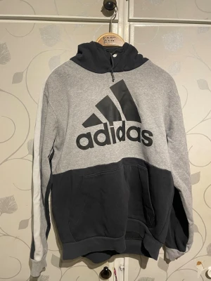 Grå och svart hoodie från Adidas - Snygg hoodie från Adidas i grått och svart med stor logga framtill. Tröjan har huva med dragsko och en stor magficka. Tillverkad i mjukt bomullsmaterial, perfekt för en avslappnad och sportig stil.